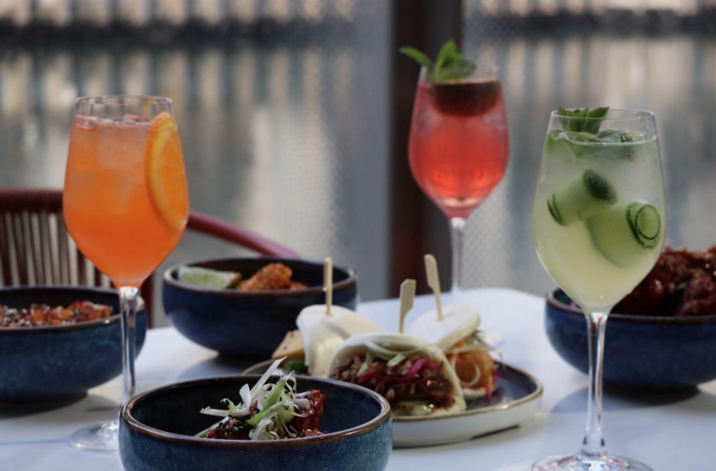 Three summer cocktails including an Aperol Spritz, pink Spritz and Eden Spritz served alongside Pan-Asian sharing plates and bao buns on the waterside terrace at Amazing Grace Canary Wharf