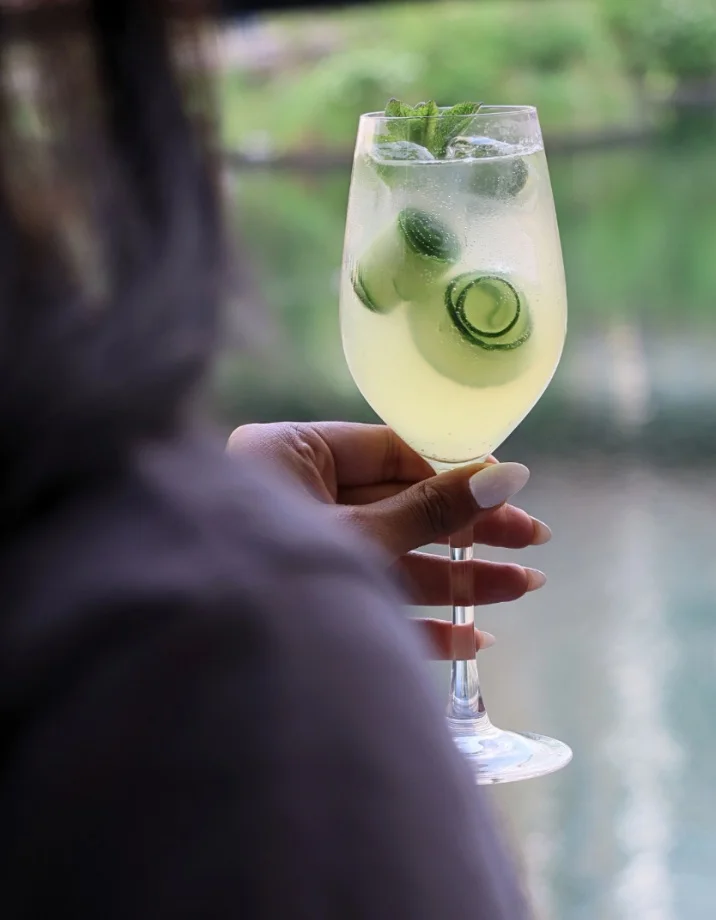 A hand holding the Eden Spritz cocktail — St-Germain, prosecco, cucumber and mint — at Amazing Grace Canary Wharf with the waterside terrace visible in the background